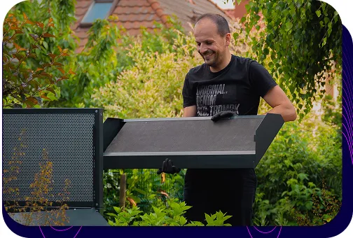 Ein lächelnder Mann, Mitarbeiter der Firma Silencions, montiert eine Verkleidung für eine Wärmepumpe im Garten eines Privathauses.