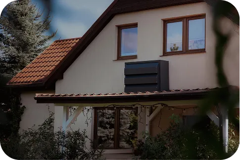 Ein Einfamilienhaus mit beiger Fassade und orangefarbenem Dach. An der Gebäudewand ist eine dunkle Silencions s|AC|-Schallschutzhaube angebracht, die den Geräuschpegel der Klimaanlage effektiv reduziert und die Ästhetik verbessert.