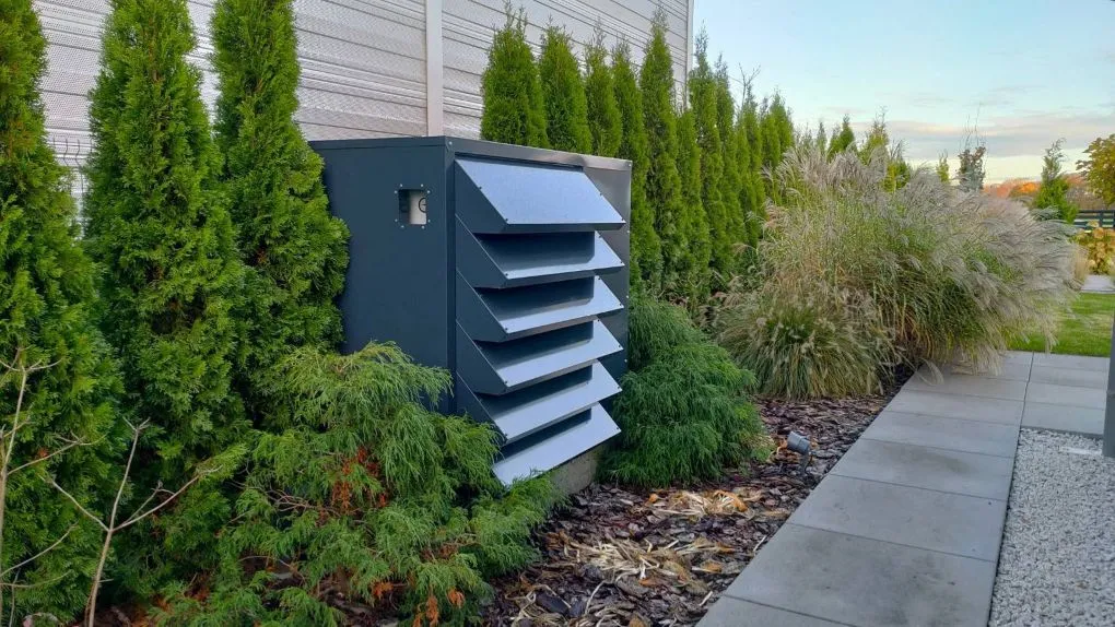 Modernes, dunkelgraues schallschutzhaube nach Maß für einen Kaltwasserkühler, ästhetisch in einen gepflegten Garten mit einer Reihe von Thujen integriert.