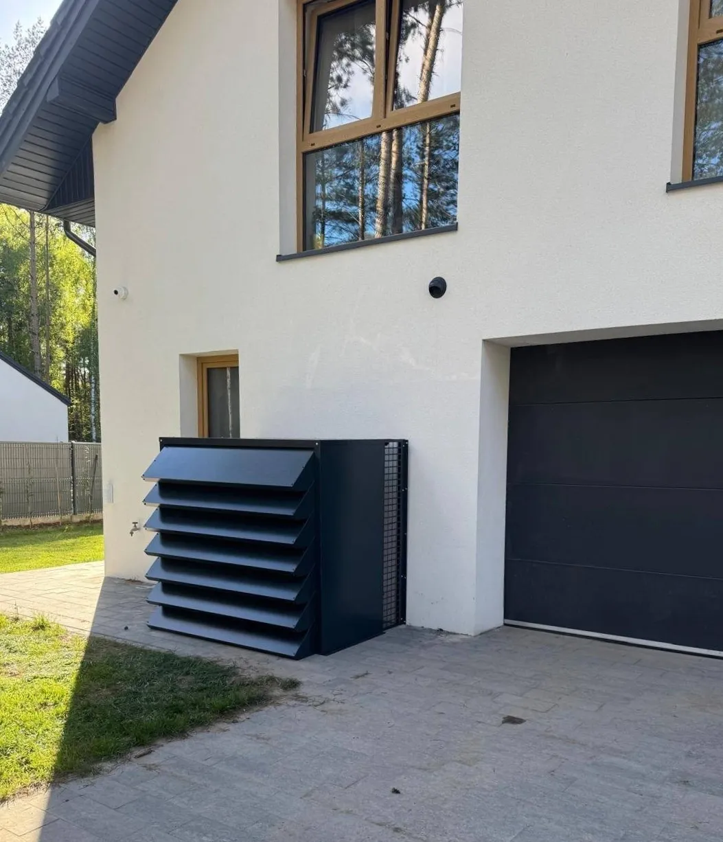 Dunkelgraues, personalisiertes schallschutzhaube für wärmepumpe, das perfekt zur weißen Fassade und zum dunklen Garagentor eines modernen Hauses passt.