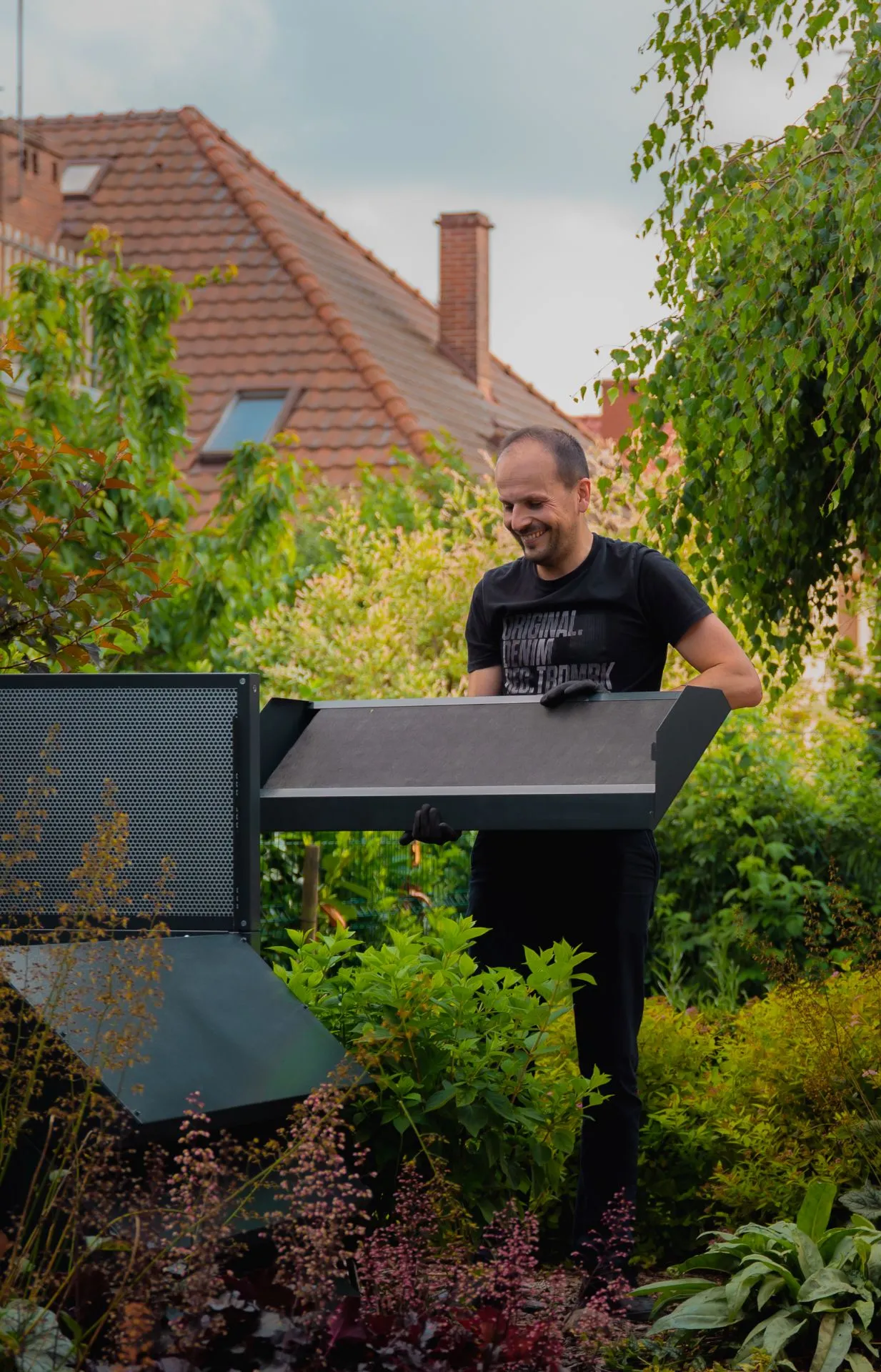 Ein lächelnder Mann, Mitarbeiter der Firma Silencions, montiert eine Verkleidung für eine Wärmepumpe im Garten eines Privathauses.