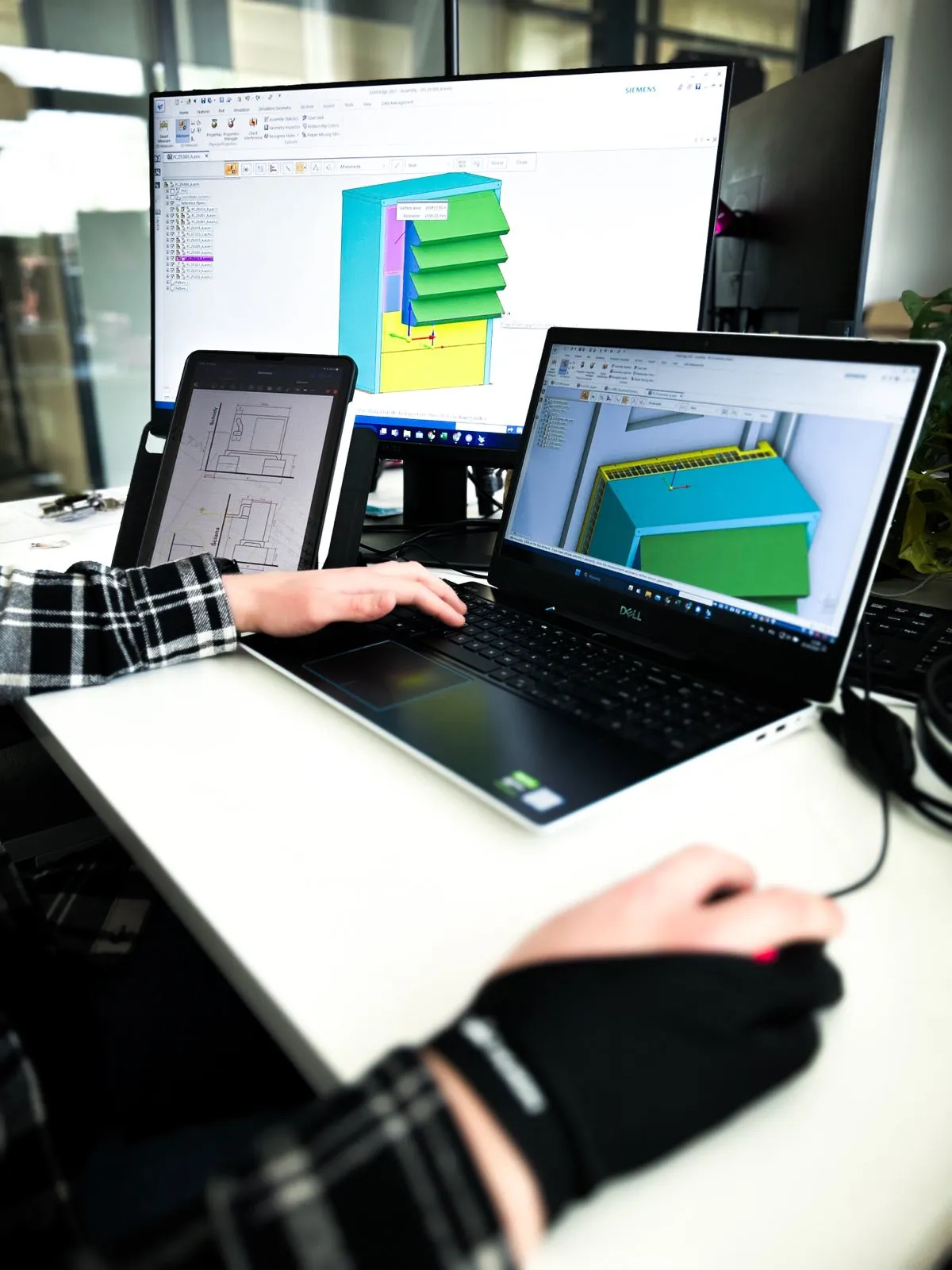 Der Ingenieur Silencions arbeitet im Rahmen des Programms an einem Projekt für ein Gehäuse für eine Wärmepumpe; auf dem Laptop-Bildschirm und dem Monitor sind ein 3D-Modell und eine technische Zeichnung zu sehen.